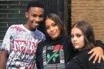 Three individuals standing close together, smiling and posing for a photo. They are dressed casually, with one wearing a graphic hoodie.