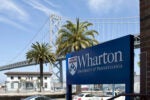 A sign for Wharton University of Pennsylvania in front of palm trees and a suspension bridge under a clear blue sky.