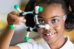 A child wearing safety goggles is holding a molecular model, smiling and examining it closely. This represents an interest in science and learning.