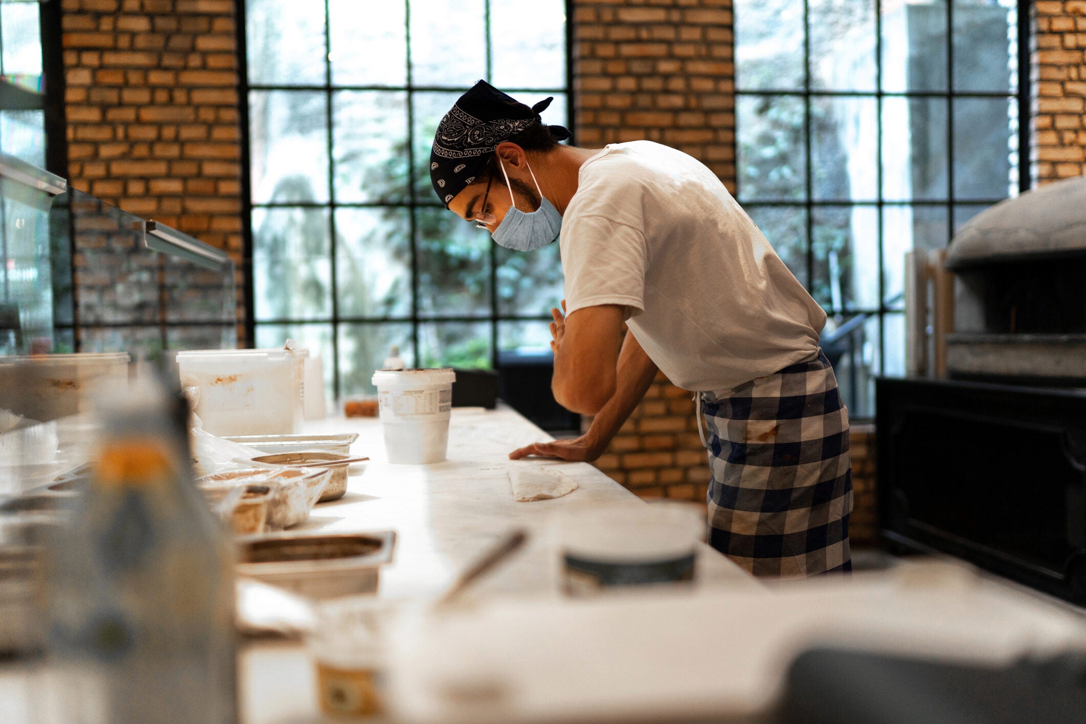 A person in a mask and bandana is preparing dough on a countertop in a bakery or kitchen setting. There are containers and ingredients on the table.