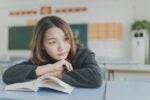 A person resting their head on folded arms on a desk with an open book in a classroom setting, appearing thoughtful or daydreaming.