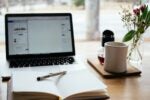 A cozy workspace with a laptop, an open notebook with a pen, a coffee mug, a small pot of coffee, and a vase of flowers on a wooden table.