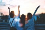 Three people standing with their backs to the camera, raising their arms in the air as they watch a sunset.