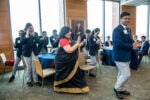 A group of people in business attire and an individual in a sari are clapping and smiling inside a conference room with tables and chairs.