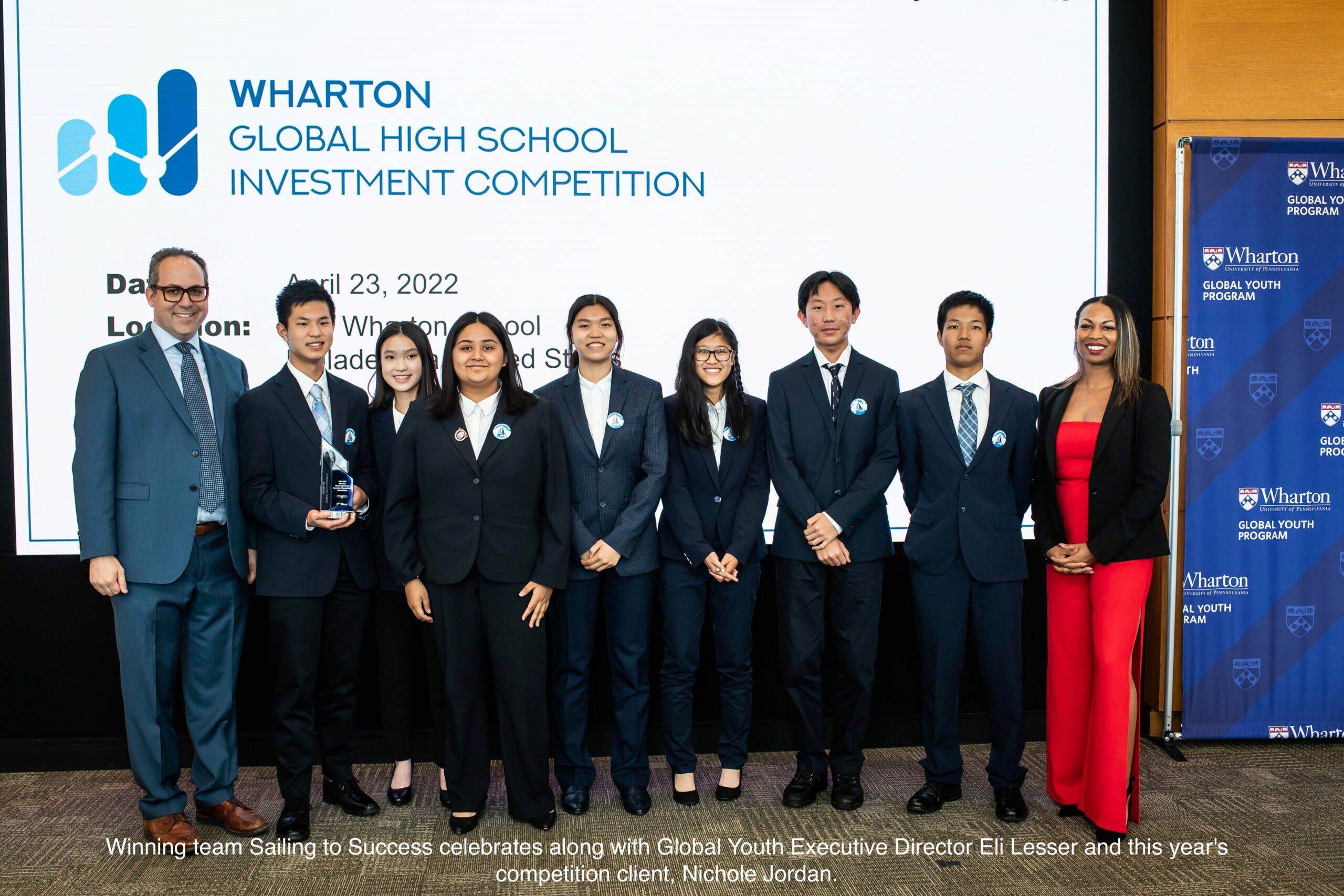 A group of individuals in formal attire stands in front of a screen displaying "Wharton Global High School Investment Competition." One person holds an award.