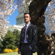 A person in a suit stands near a blossoming cherry tree, with the US Capitol building in the background.