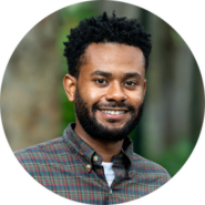 Headshot of a person with a beard wearing a plaid shirt, smiling outdoors.