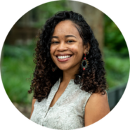 A headshot of a person with curly hair, smiling and wearing a sleeveless patterned top and earrings. The background is blurred greenery.