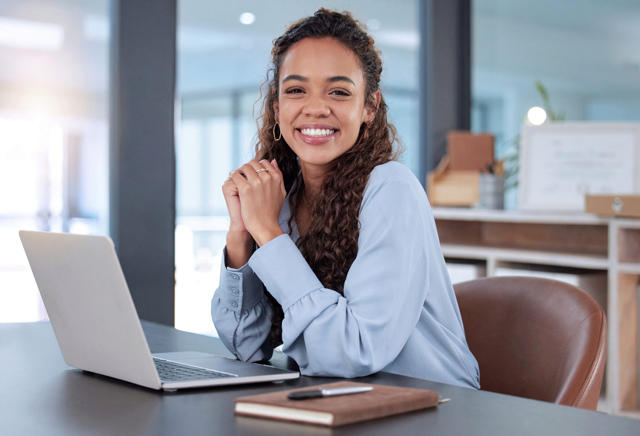 A person with curly hair smiling, sitting at a desk with a laptop and notebook in an office setting.