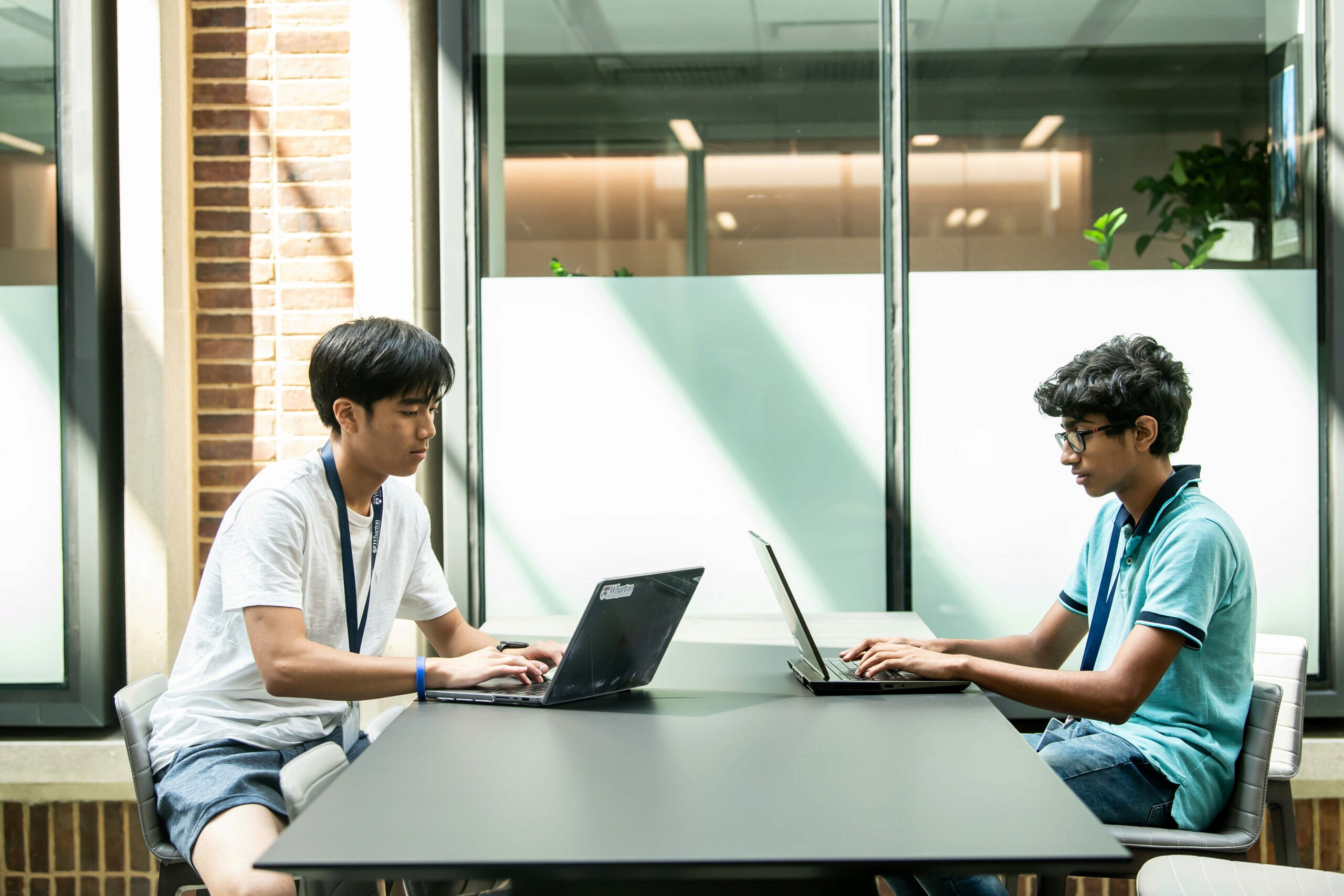 220715 KWHS GYLA 0988 Two individuals sitting across from each other at a table, using laptops. The setting appears to be a modern indoor space with natural light.