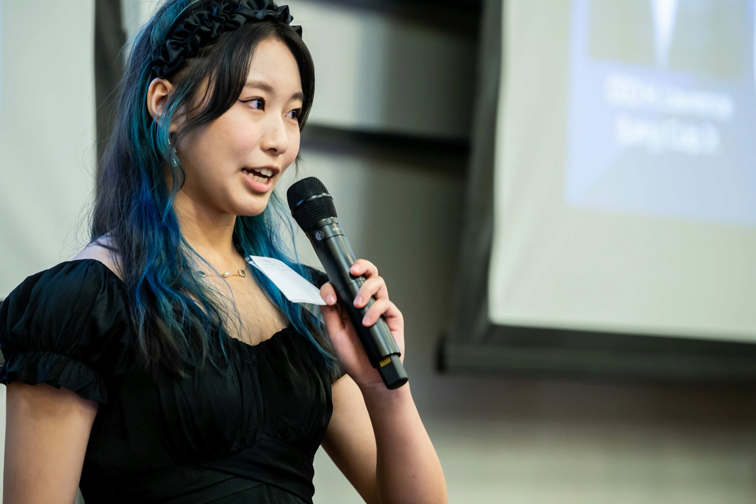 A person with long hair, wearing a black dress, holding a microphone and speaking, with a presentation screen in the background.