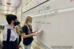 Students wearing masks work on financial equations at a whiteboard in a classroom. The equations involve present value and discount rates.