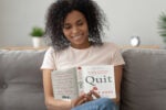 A person sitting on a couch, smiling while reading the book "Quit" by Annie Duke. A plant and a clock are in the background.