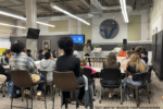 A person standing in front of an audience giving a presentation in a modern classroom or meeting space, with a screen and tables.