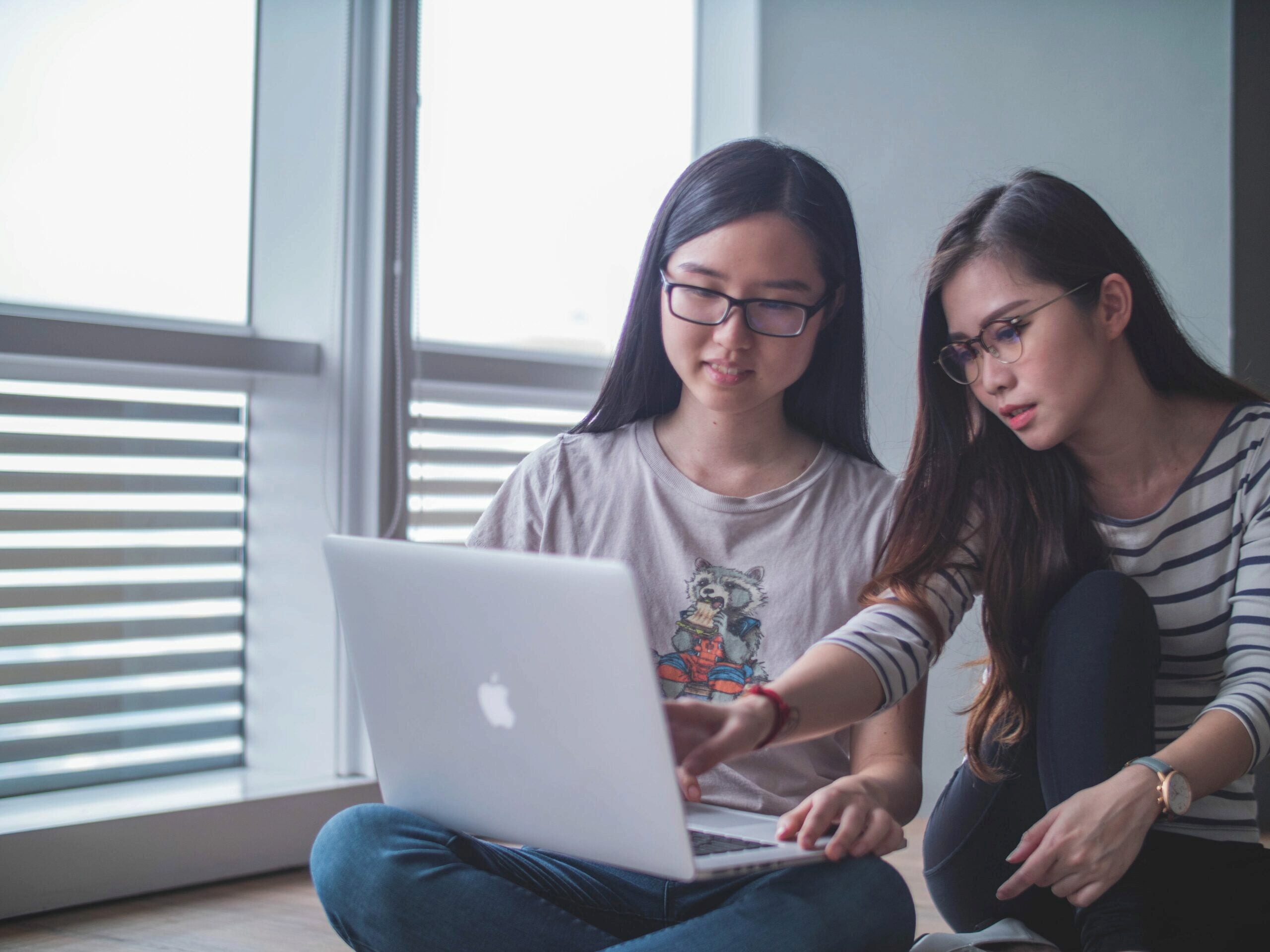 Two people are sitting on the floor, focused on a laptop. One points at the screen, suggesting collaboration or learning. They are near a window.