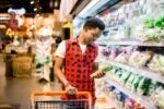 A person is shopping in a grocery store, pushing a cart and examining a bag of fresh greens in the produce section.