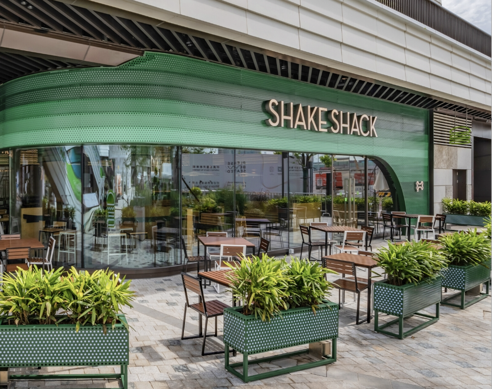 Outdoor seating area of a Shake Shack restaurant with green planters and glass windows. The sign reads "Shake Shack."