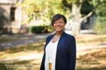 A person standing outdoors, smiling, wearing a blue jacket and white top. Surroundings include trees and sunlight. Text reads "Wharton''s first Chief Diversity, Equity & Inclusion Officer, Renita Miller."