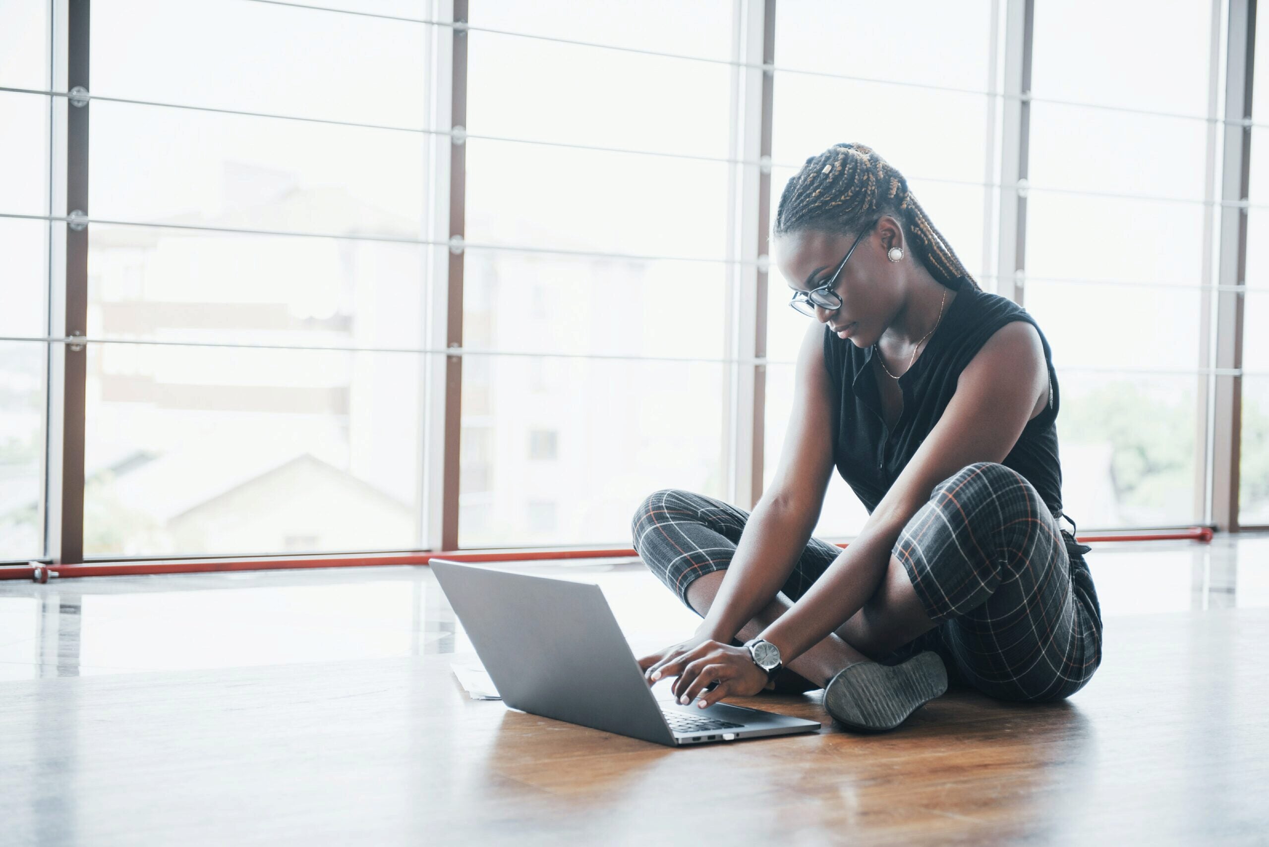 A person with braided hair and glasses sits cross-legged on the floor using a laptop near a large window.