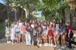 A group of young people standing together outdoors on a sunny day, posing for a photo in front of a leafy background.