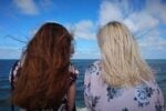 Two people with long hair facing the ocean on a windy day, with a bright blue sky and scattered clouds in the background.