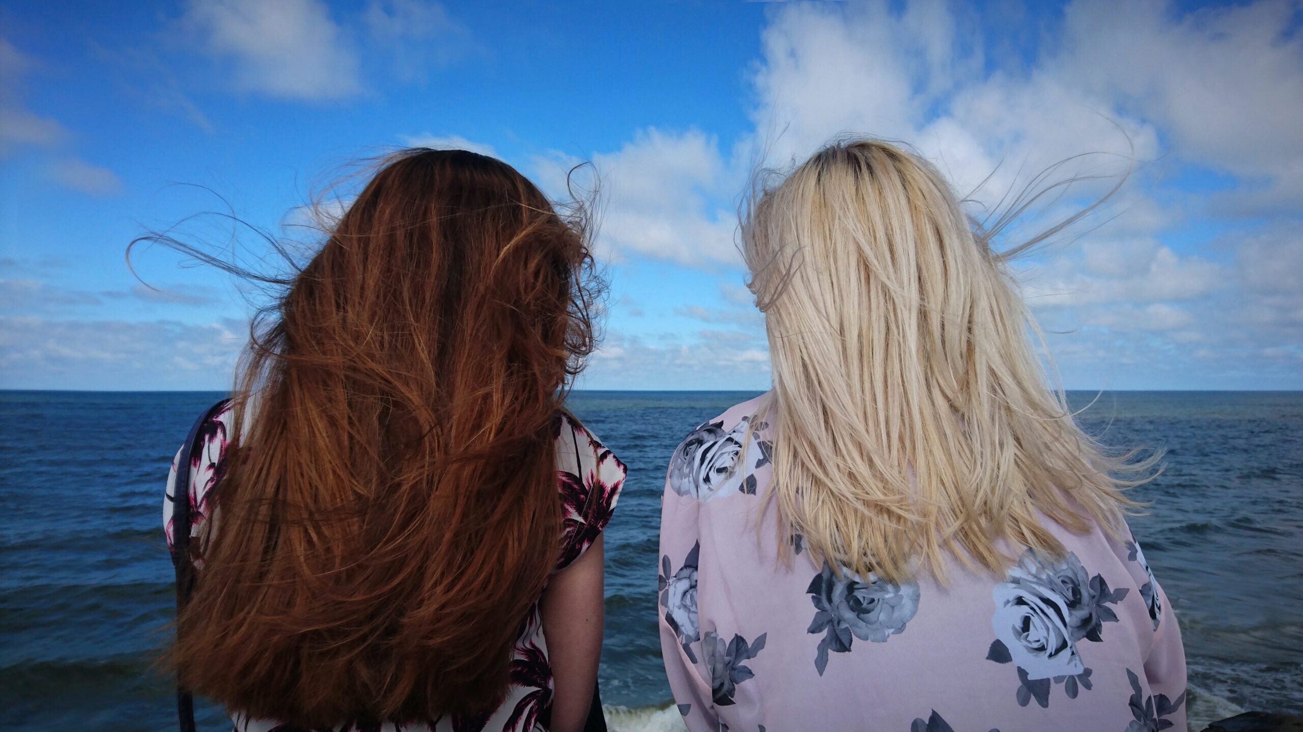 Two people with long hair facing the ocean on a windy day, with a bright blue sky and scattered clouds in the background.