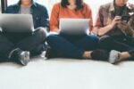 Three people sitting on the floor, two using laptops and one using a smartphone.