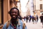 A person smiling, wearing a denim jacket and a backpack, standing on a busy city street with blurred pedestrians in the background.