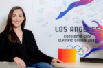 A person posing for a headshot beside a colorful sculpture and a wall with signatures. The poster in the background reads "Los Angeles Candidate City Olympic Games 2024" with Olympic rings.