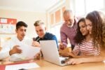 A group of students and a teacher are working together at a table, focusing on a laptop and documents. The setting is a classroom.