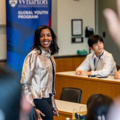 A person standing in a classroom, smiling, with a "Wharton Global Youth Program" banner in the background. Students are seated at desks listening.