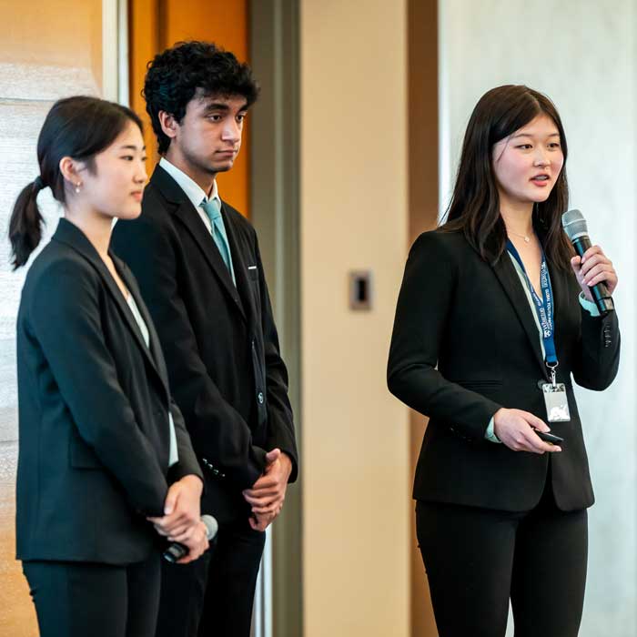 Three individuals in business attire are presenting. One person is speaking into a microphone, while the other two are attentively observing.