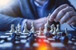 A person playing chess, focusing on moving a white piece on a chessboard. The image represents strategy and critical thinking.