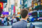 Person in a suit with a shoulder bag standing on a busy city street, surrounded by pedestrians and blurred bright signs.