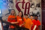 A small group of people wearing orange shirts stands on a staircase in front of a wall featuring sports and school-themed images.