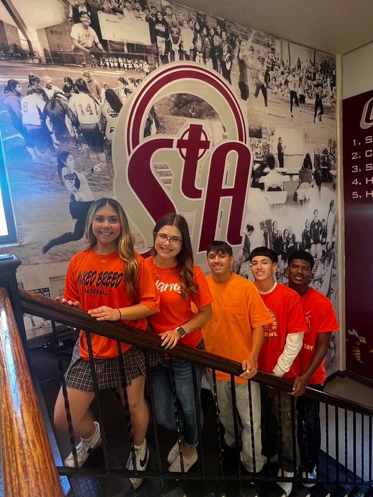 A small group of people wearing orange shirts stands on a staircase in front of a wall featuring sports and school-themed images.