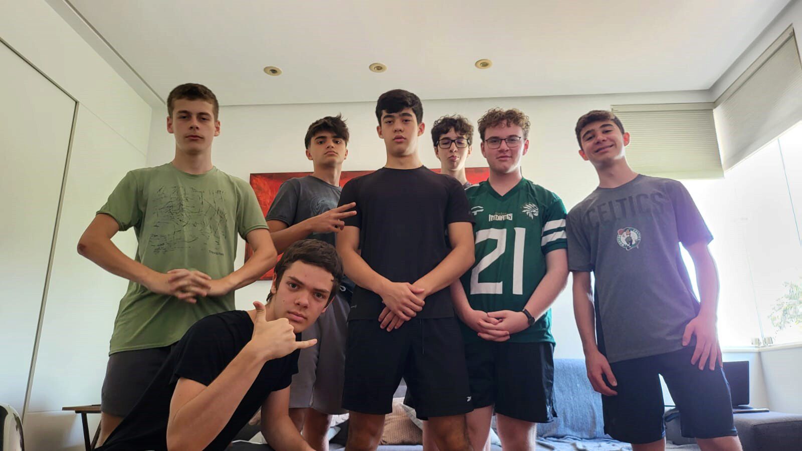 A group of young people poses indoors, some with casual gestures. They are wearing a mix of athletic and casual clothing. The room has modern decor.