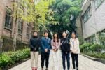 A group of five people standing outdoors on a pathway between two brick buildings, surrounded by greenery. They are dressed casually, with trees and plants visible in the background.
