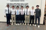 A group of young people standing side by side, dressed in formal and semi-formal attire. They are smiling, and a wall with text serves as a backdrop.