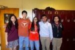 A group of five people standing in front of a row of red lockers. They are smiling and posing casually, suggesting a school environment.