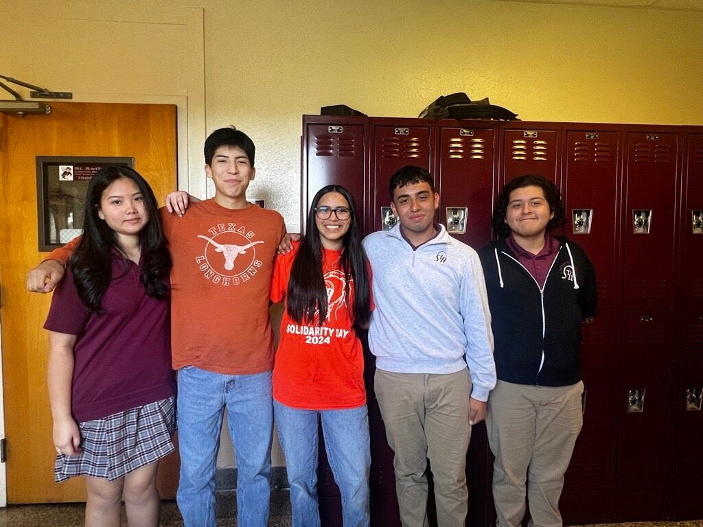 A group of five people standing in front of a row of red lockers. They are smiling and posing casually, suggesting a school environment.