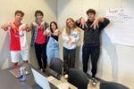 A group of five people posing with hand signs in a casual office setting. A laptop and drinks are on the table, and a poster titled "Tripple M" is on the wall.