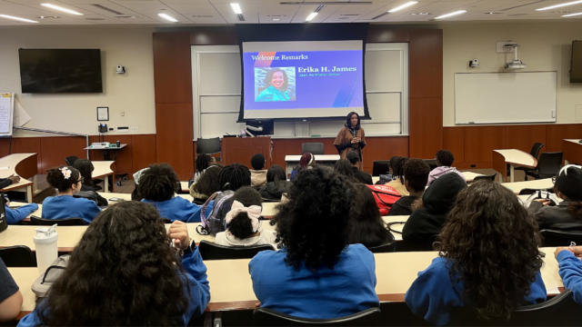 A person giving a lecture in a classroom setting with a presentation slide displaying a headshot and the name "Erika H. James, Dean, the Wharton School." Students are seated, listening attentively.