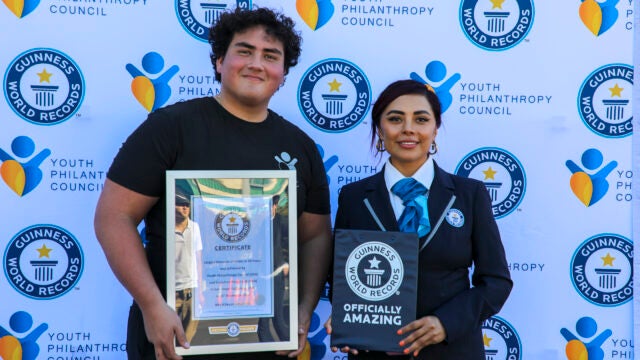 A small group celebrating a Guinness World Record achievement with a certificate. They are standing in front of a backdrop displaying logos for the Youth Philanthropy Council and Guinness World Records.