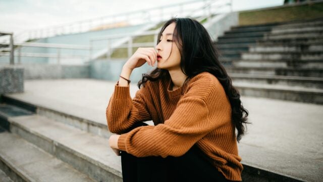 A person is sitting on outdoor steps, wearing a brown sweater and black pants. They appear to be in a thoughtful pose, with their hand near their face, against a background of gray steps and railings.