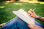 A person writing in a notebook with a pen while sitting on the grass, wearing a bracelet and jeans. This represents relaxation, creativity, or journaling outdoors.