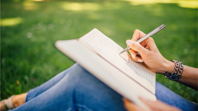 A person writing in a notebook with a pen while sitting on the grass, wearing a bracelet and jeans. This represents relaxation, creativity, or journaling outdoors.