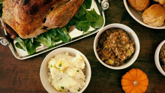 A festive meal featuring a roast turkey on a platter with greens, mashed potatoes, stuffing, rolls, and a small decorative pumpkin.