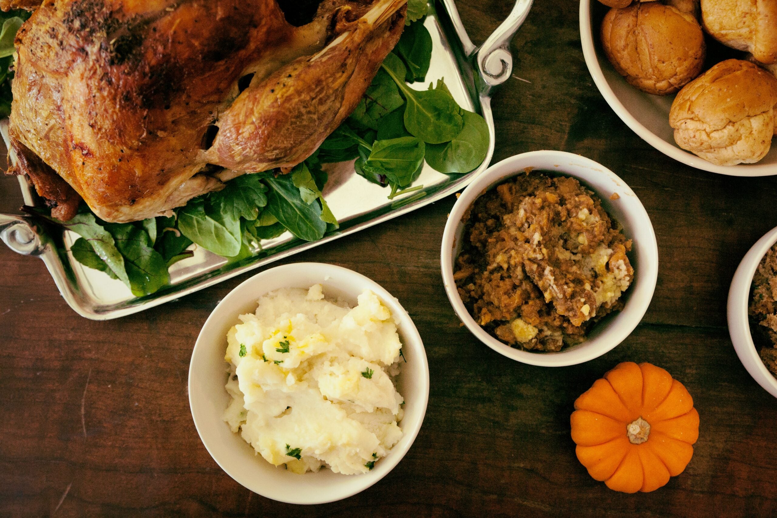 A festive meal featuring a roast turkey on a platter with greens, mashed potatoes, stuffing, rolls, and a small decorative pumpkin.