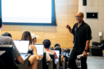 A smiling professor dressed in black talks to a lecture hall filled with high school students